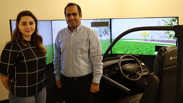 A professor and graduate assistant stand in front of a driving simulator
